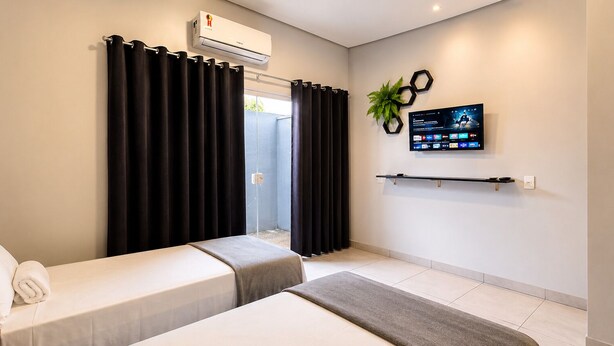 Quarto de hotel com cama, uma pequena mesa, TV fixada na parede e janela com cortinas.