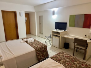 Habitación de hotel con dos camas, un escritorio con computadora, una silla, un televisor y un refrigerador pequeño.