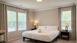 A bedroom with a bed, two bedside tables with lamps, a clock, and a vase with flowers. - Brand New 3BR - Patio - Fast WiFi+Workspace - Near Airport & Whitewater Center (Charlotte)