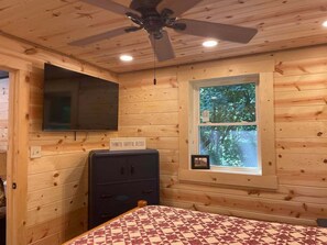 Habitación tipo cabaña de madera con cama, ventilador de techo, televisor montado en la pared, ventana con vistas a árboles y aparador con cajones.