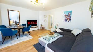 A modern living room with a flat-screen TV, a sofa, a glass coffee table, and a dining area with blue chairs. - Charmante Wohnung in Koblenz (Koblenz)