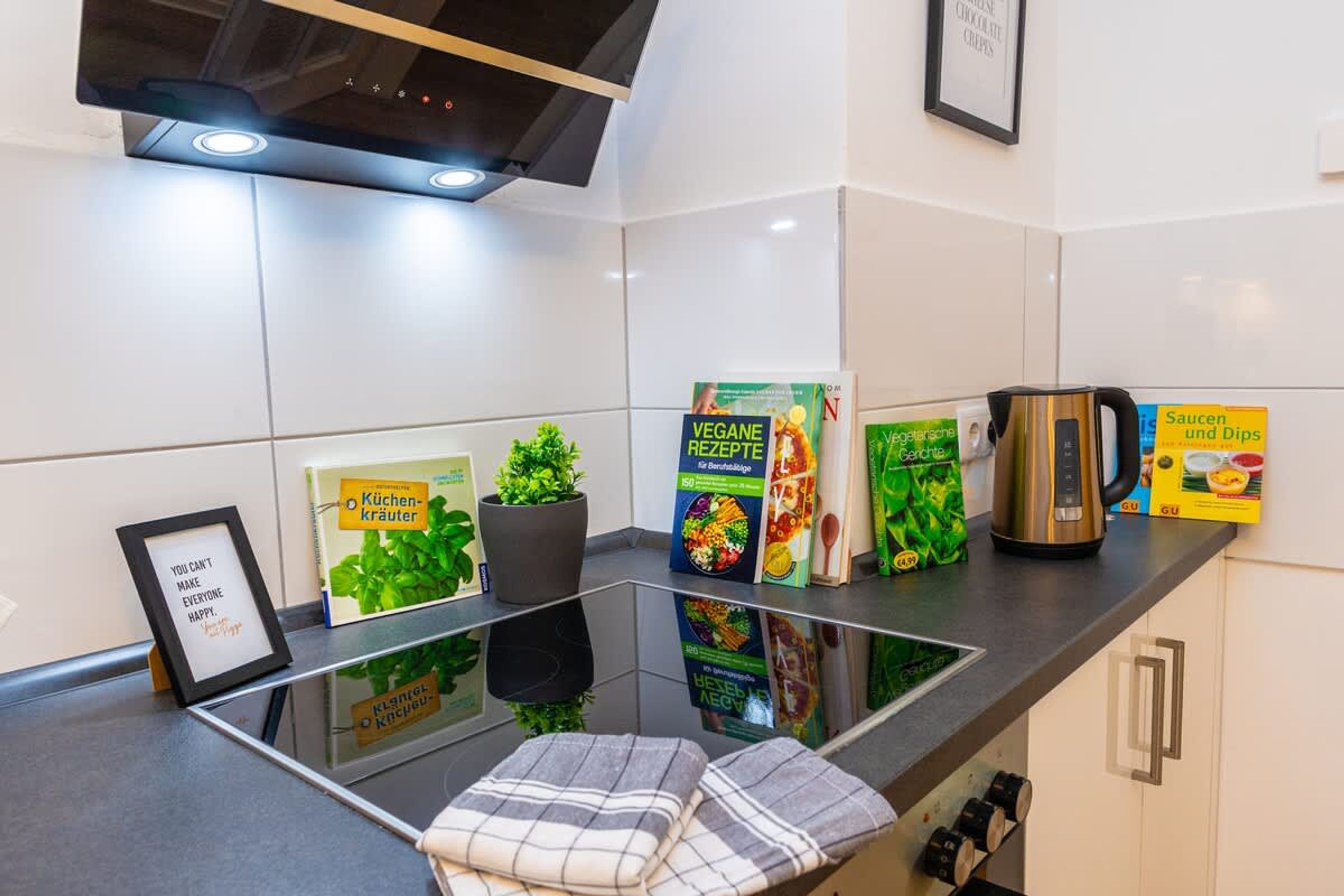 A modern kitchen with a glass countertop, a kettle, a framed quote, and various cookbooks.