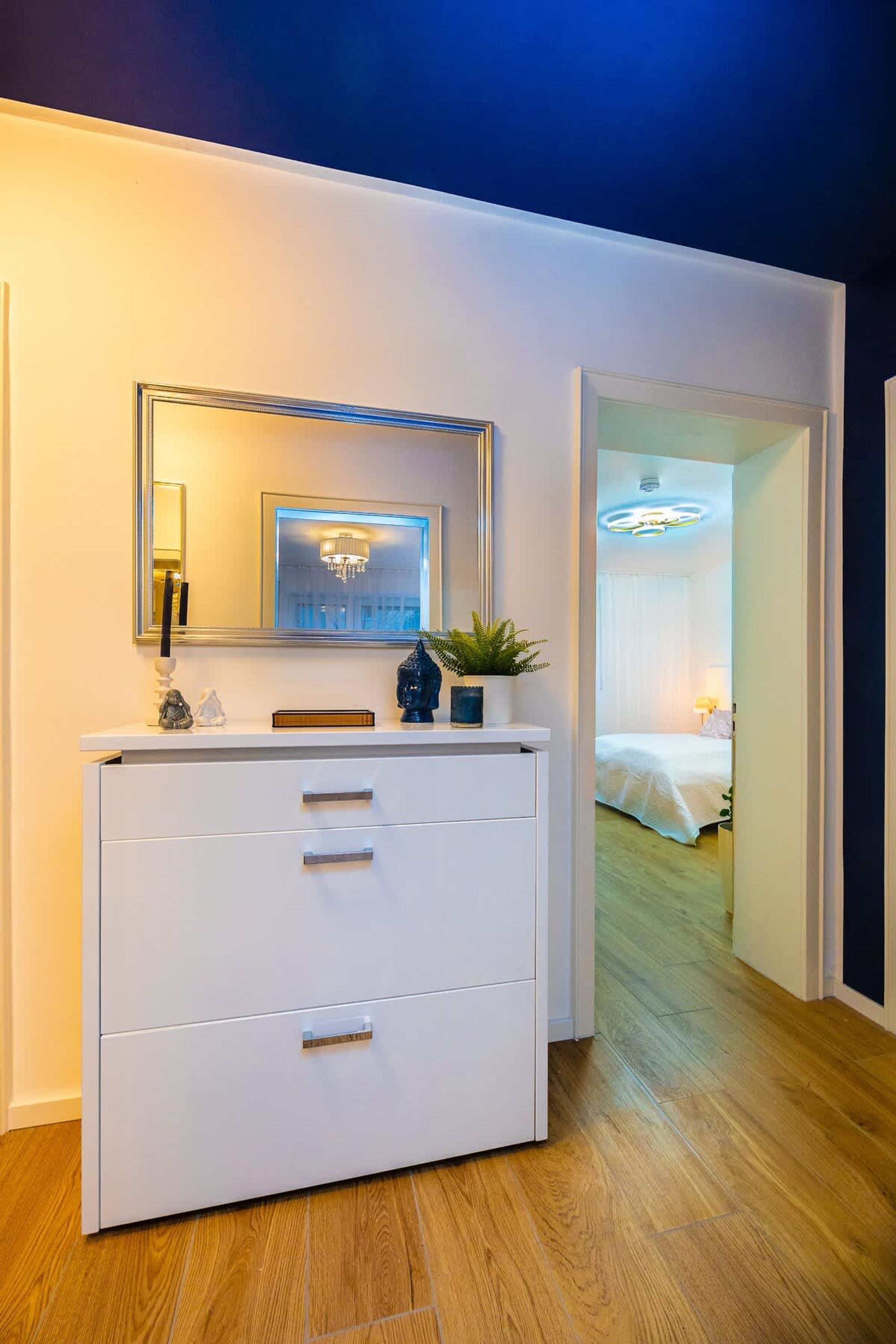 Une chambre d’hôtel moderne avec une commode blanche, un miroir, une plante en pot et une vue sur la chambre adjacente.