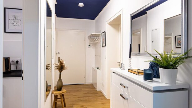 A hotel corridor with a wooden floor, white walls, a ceiling light, a mirror, a plant, and a small stool. - Charmante Wohnung in Koblenz (Koblenz)