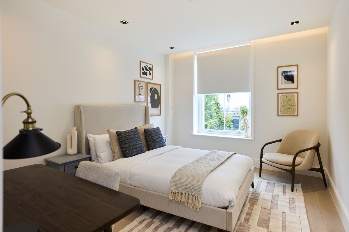 A bedroom with a bed, bedside table, lamp, chair, and artwork on the walls.