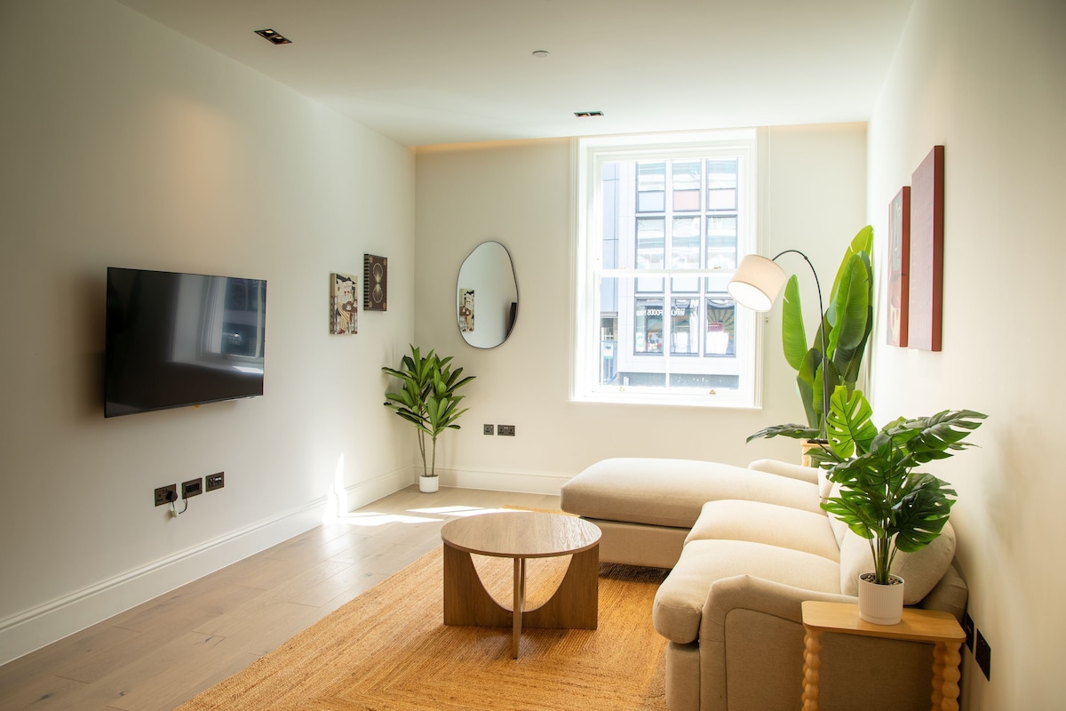A modern living room with a flat-screen TV, a round coffee table, a beige sofa, a large mirror, and a potted plant.