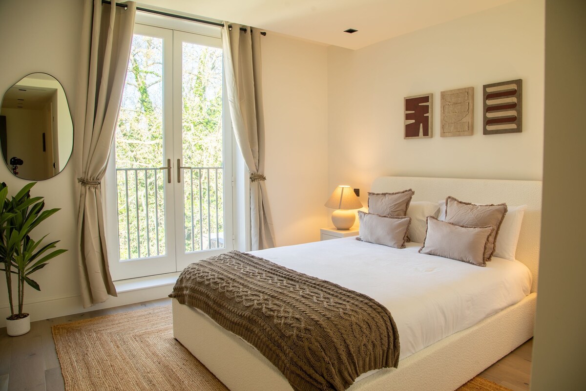 A bedroom with a bed, curtains, a bedside lamp, a mirror, and a potted plant.