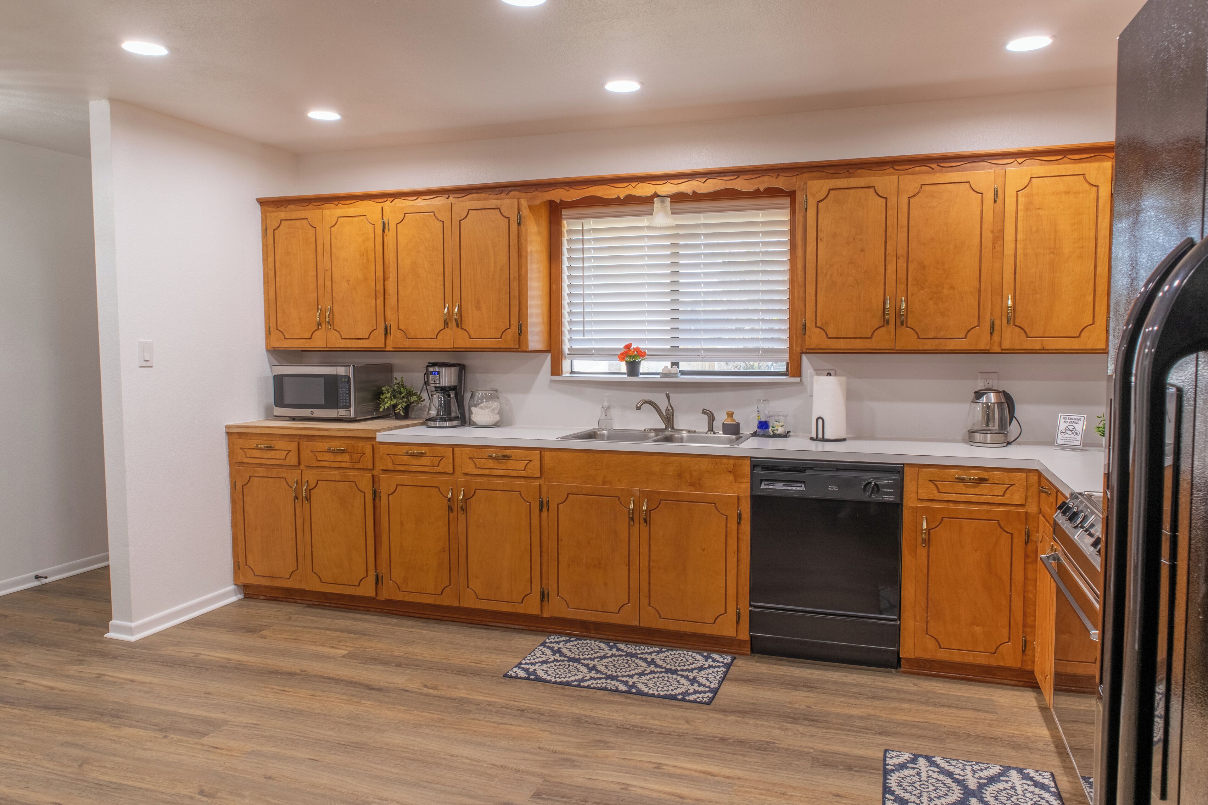 Una cocina con armarios de madera, microondas, lavavajillas y una ventana con persianas.