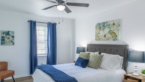 Ein Schlafzimmer mit Bett, Nachttisch, Sessel, Deckenventilator und einem Fenster mit Vorhängen.