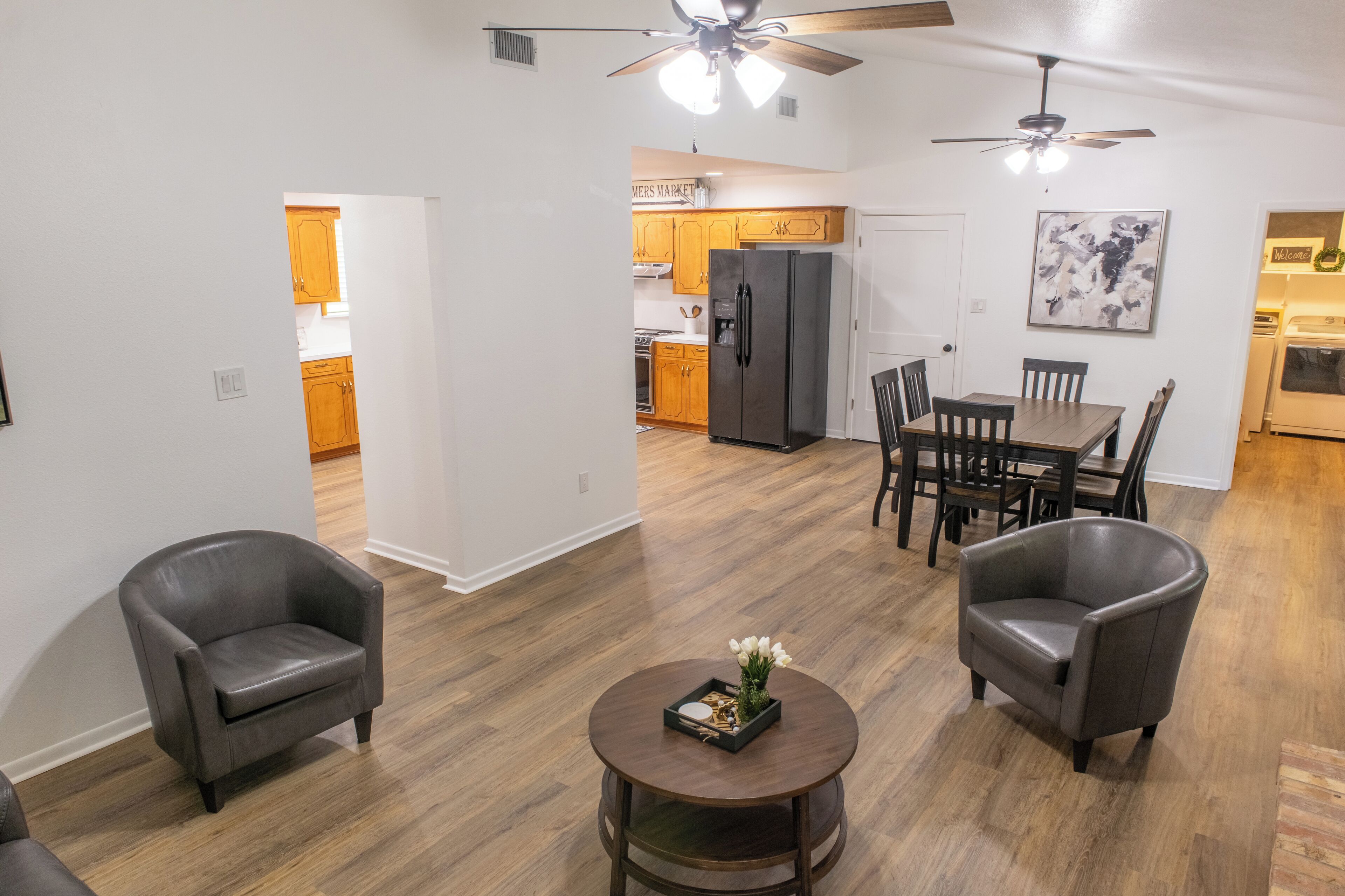 A modern living room with a dining area, two leather armchairs, a round coffee table, and a ceiling fan.
