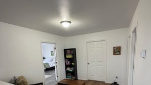 Ein Zimmer mit weißer Decke, Holzboden, einem kleinen Tisch mit Stuhl, einem Bücherregal und einer Tür.