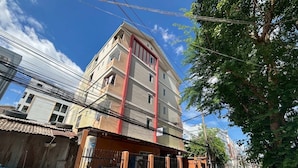 A multi-story building with a gated entrance, a visible trash bin, and a tree in the foreground. - 401 Budget Room Utilities Wifi Balcony in Ratchada (Bangkok)