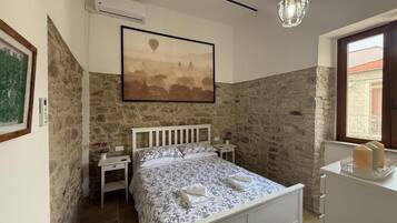 A bedroom with a stone wall, a bed with a floral bedspread, a bedside table, a nightstand, a chandelier, and a window with a view.