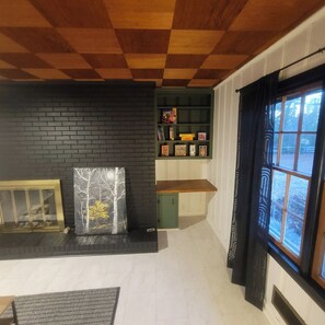 A room with a black brick wall, a wooden ceiling, a bookshelf, a framed picture, and a window with curtains. - Raleigh Urban Cabin in the Woods, Pet Friendly, Covered Carport (Raleigh)