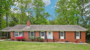 A single-story brick house with a front porch, a chimney, and a small garden with pink flowers. - Raleigh Urban Cabin in the Woods, Pet Friendly, Covered Carport (Raleigh)