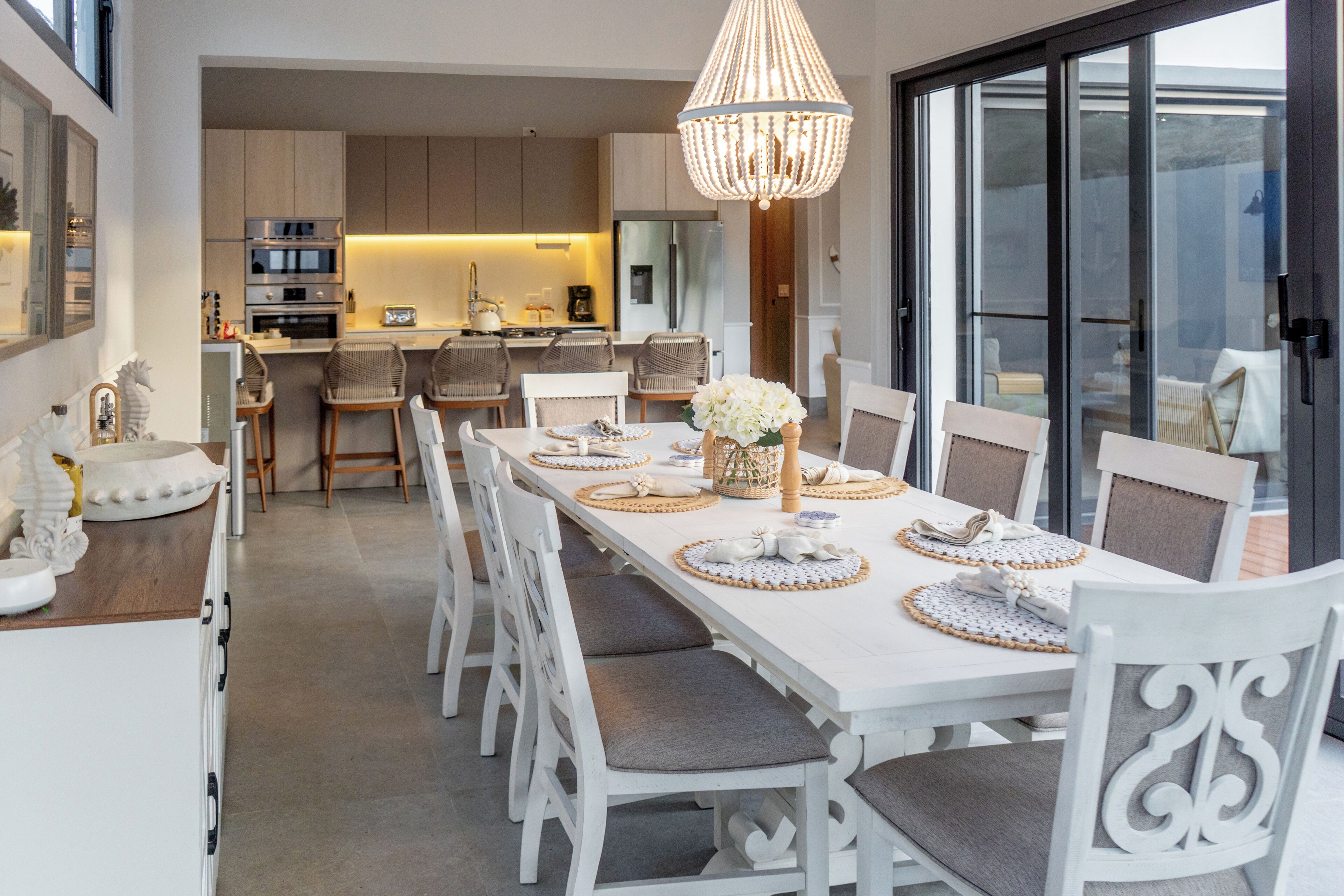 Un comedor con una larga mesa, sillas blancas y una araña.
