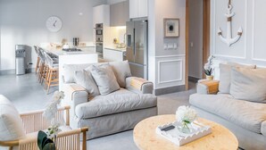 Ein modernes Wohnzimmer mit Sofa, Sesseln, Couchtisch und einer Küchenzeile inklusive Kühlschrank und Esstisch.