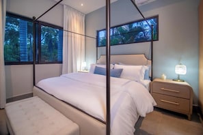 A bedroom with a canopy bed, a bedside table with a lamp, and a view of trees through the window.