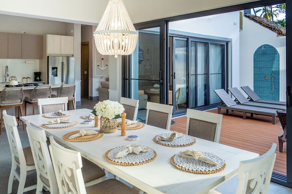 A dining area with a white table set for six, surrounded by white chairs, a chandelier, and a view of a pool and lounge chairs.