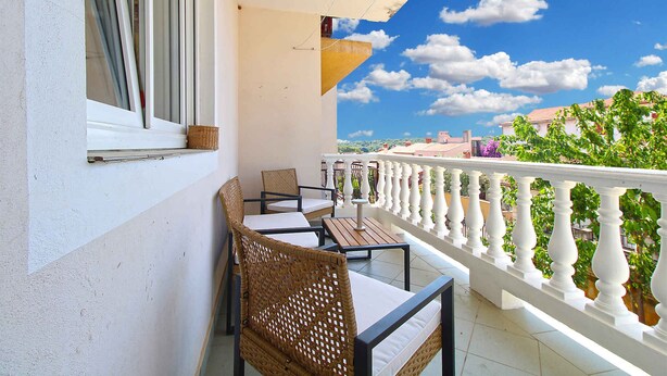 Ein Balkon mit Korbmöbeln, einer weißen Brüstung und Blick auf Gebäude und Grünflächen.