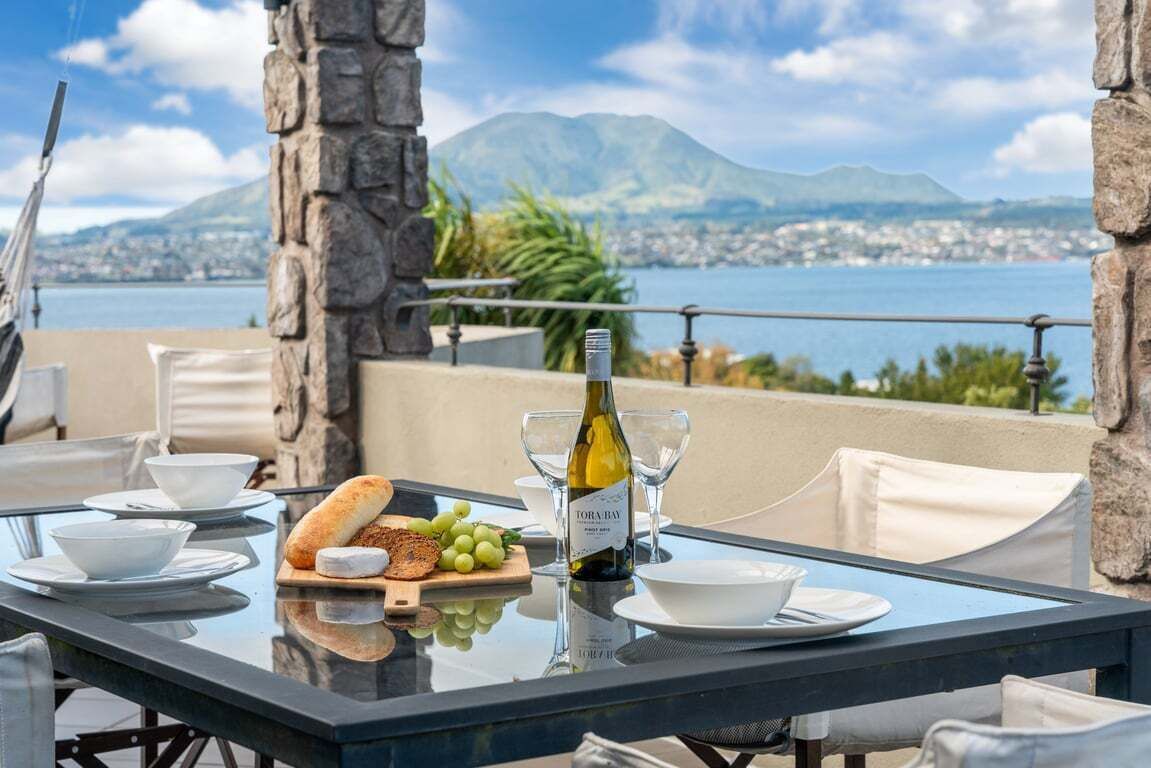 A table set for dining with a bottle of wine, bread, and grapes, overlooking a scenic view of a lake and mountains.
