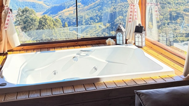 A wooden cabin with a large window, a hot tub, and a view of a mountainous landscape. - Country house in the mountains of Santa Catarina, at 1000m altitude, surrounded by nature. (Alfredo Wagner)