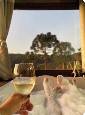 A person holding a glass of white wine, a bathtub with bubbles, and a view of trees through a window. - Country house in the mountains of Santa Catarina, at 1000m altitude, surrounded by nature. (Alfredo Wagner)