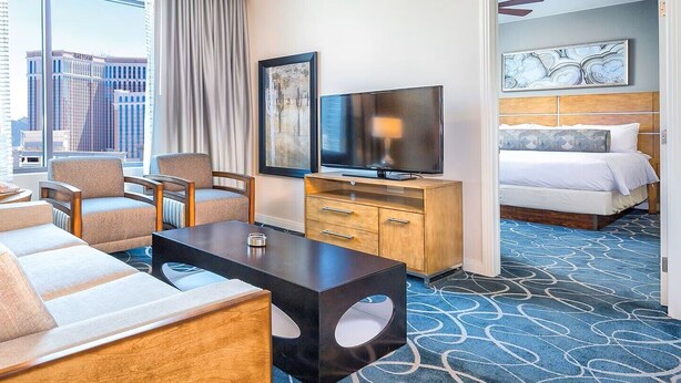 Et hotelværelse med sofa, stole, et fjernsyn, en seng og udsigt til bygninger.