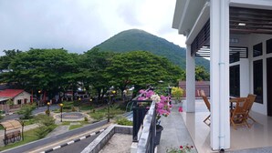 A balcony with a view of a mountain, a road with a roundabout, and a building with a white facade. - D' Kabong SIRIH Homestay (Bandanaira)