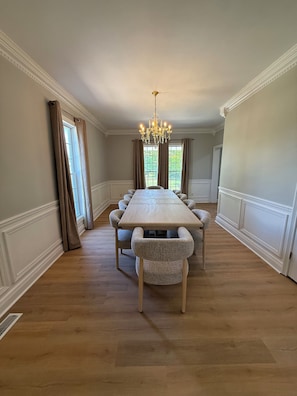 Ein Esszimmer mit einem langen Tisch, Stühlen, einer Chandelier und großen Fenstern mit Vorhängen.