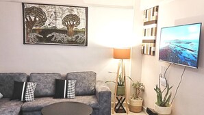Ein Wohnzimmer mit grauem Sofa, einem kleinen runden Tisch, einer Stehlampe, einem Fernseher und einem Gemälde.