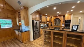 A modern kitchen with wooden cabinetry, a stainless steel refrigerator, and a large island with glass doors. - Spacious Modern Home on Acreage (Melissa)