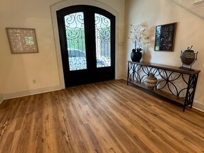 Een gang met een houten vloer, een zwarte deur met decoratieve glaspanelen, een console tafel met vazen, en twee ingelijste kunstwerken aan de muur.
