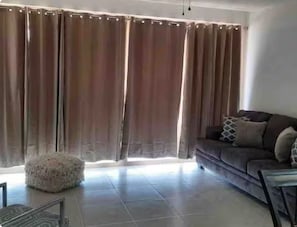 Ein Wohnzimmer mit einer Couch, einem kuscheligen Ottomanen und einem großen Fenster mit Vorhängen.