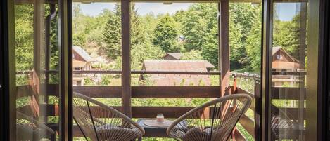 A balcony with two wicker chairs and a table, overlooking a forested area with houses.