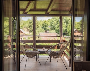 Un balcón con sillas de mimbre y una mesa, con vistas a una zona boscosa con casas.