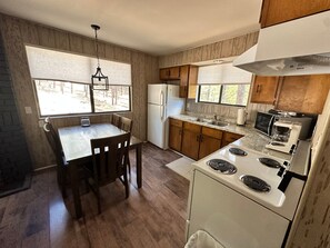 A compact kitchen with wooden cabinets, a stove, a refrigerator, and a dining table with chairs. - Cozy Cabin in Greer: Hike, Bike & Ski! (Greer)