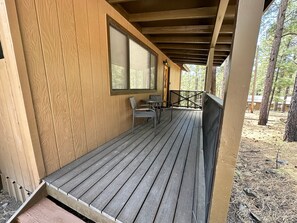 A wooden deck with a table and chairs, a window, and a view of the surrounding forest. - Cozy Cabin in Greer: Hike, Bike & Ski! (Greer)