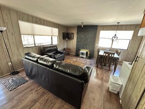 A living room with a black leather sofa, a fireplace, a television, and a dining area with a table and chairs. - Cozy Cabin in Greer: Hike, Bike & Ski! (Greer)
