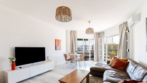 Een moderne woonkamer met een flatscreen televisie, een bank, een salontafel, pendelverlichting van riet, een eethoek met tafel en stoelen, en grote ramen met gordijnen.