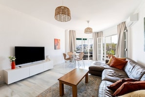 Ein modernes Wohnzimmer mit Flachbildfernseher, einer Couch, einem Couchtisch, Weiden-Pendelleuchten, einem Essbereich mit Tisch und Stühlen sowie großen Fenstern mit Vorhängen.