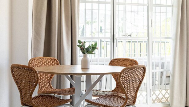 Een ronde tafel met vier rieten stoelen, een vaas met bloemen en uitzicht door een raam met witte lijst.