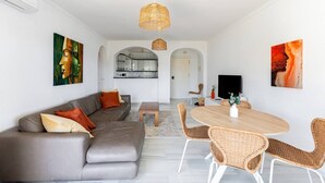 Een moderne woonkamer met een grijze bank, rieten stoelen, een houten eettafel en kunstwerken aan de muur.