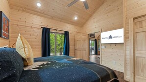 Une chambre dans un chalet en bois, avec un lit, un ventilateur de plafond, une fenêtre avec des rideaux et une porte.