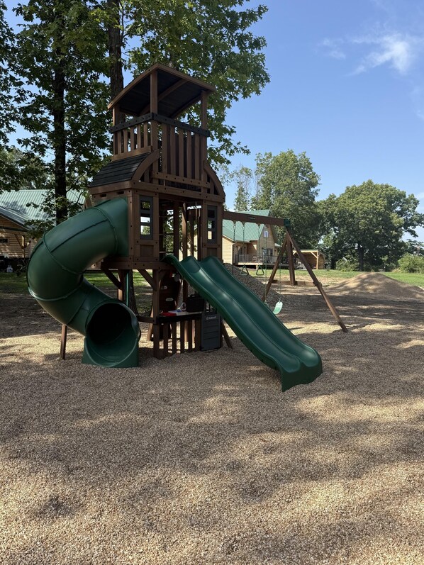 A wooden play structure with a slide and climbing area, surrounded by trees and gravel.