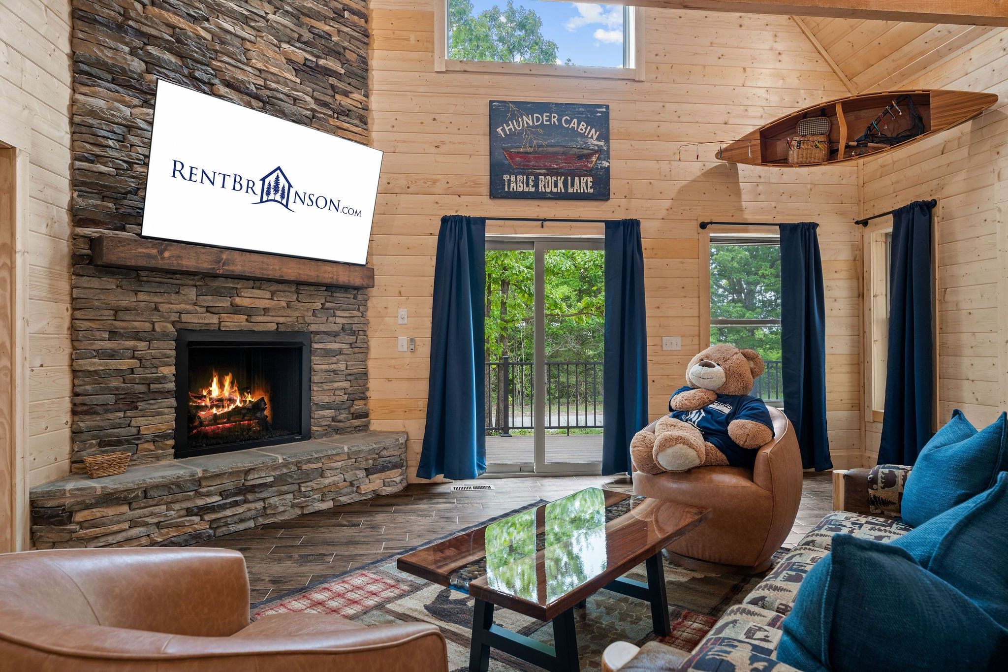 Interior de una cabaña de troncos con chimenea de piedra, un letrero que dice 'Thunder Cabin Table Rock Lake' y un oso de peluche grande en un sofá.