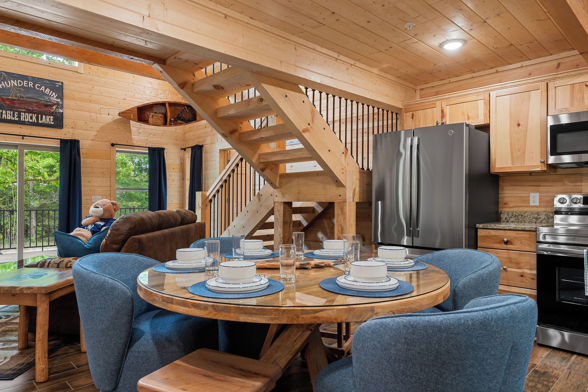 A cabin interior with a dining table set for four, a kitchen with modern appliances, and a staircase leading to an upper level.