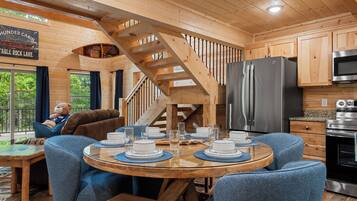 A cabin interior with a dining table set for four, a kitchen with modern appliances, and a staircase leading to an upper level.