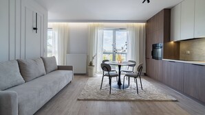 Ein modernes Wohnzimmer mit Sofa, einem Esstisch mit Stühlen, einer Küchenzeile mit Schränken und einem großen Fenster mit Vorhängen.