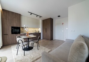 Un salon moderne avec un canapé, un coin repas comprenant des chaises et une table, une cuisine dotée d’un four encastré et un téléviseur à écran plat fixé au mur.